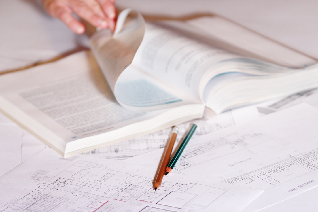 Hand turning page of book on top of blueprint drawings and pencils